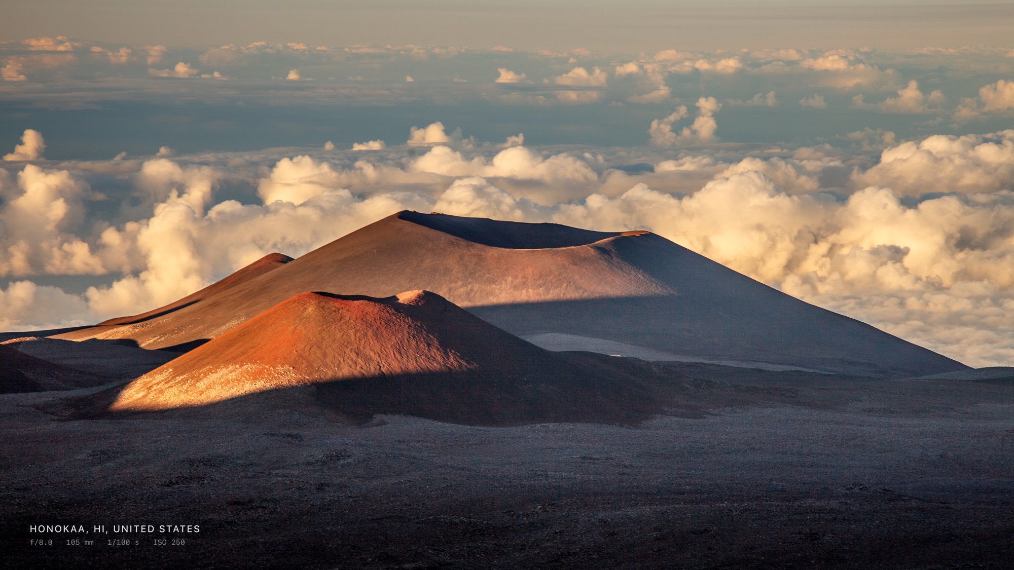 Volcanic cinder cones above a sea of clouds at sunset, filling an Apple TV screen, with the capture location and camera settings shown discreetly in the corner